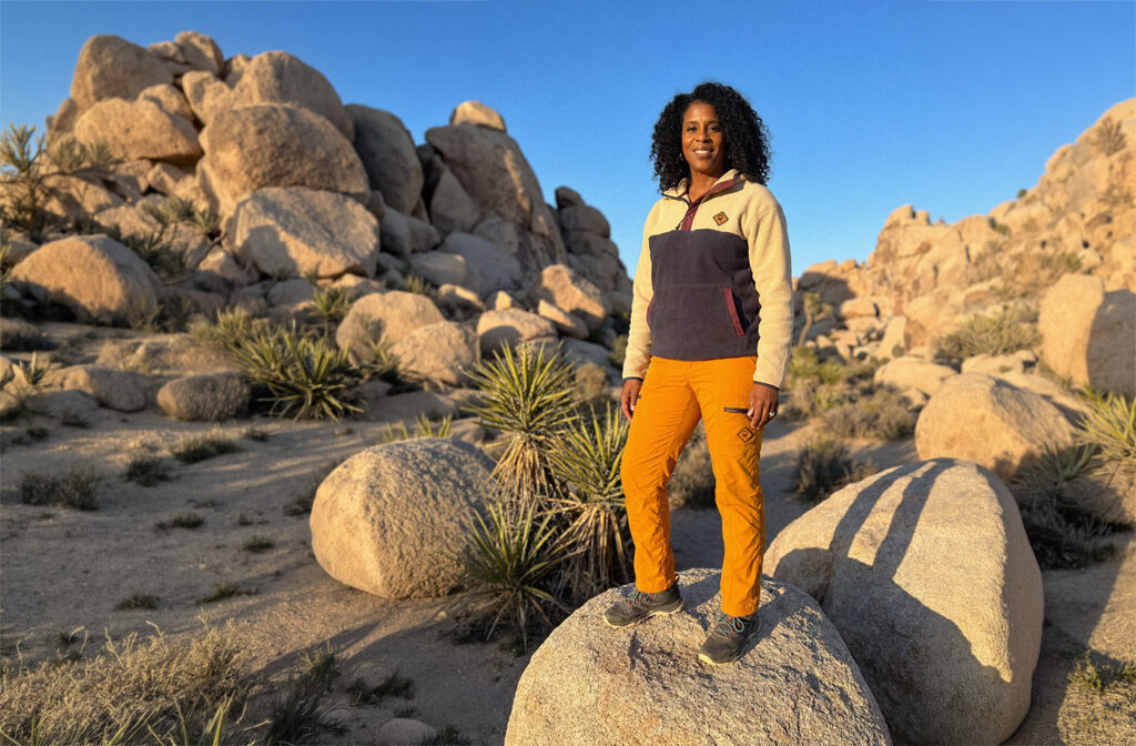 Woman standing on a large rock in a desert landscape wearing outdoor trail pants and hiking apparel during a wilderness adventure.