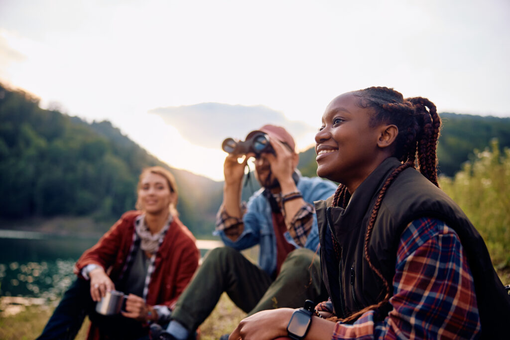 Group of friends relaxing by a mountain lake during sunset while birdwatching with binoculars in a peaceful outdoor setting.