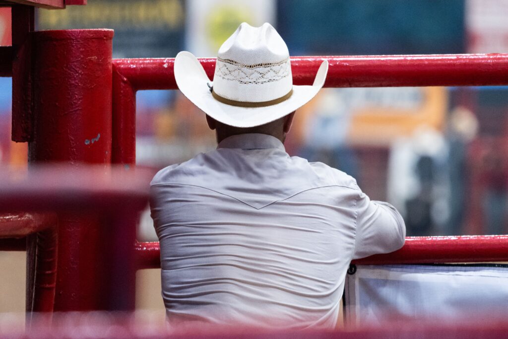 Black cowboy in a white hat leaning against a red rodeo fence, reflecting on tradition and heritage at a livestock arena.