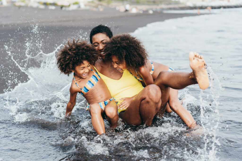 Black mother and her two daughters laughing and playing in shallow ocean waves at the beach during a family summer outing.