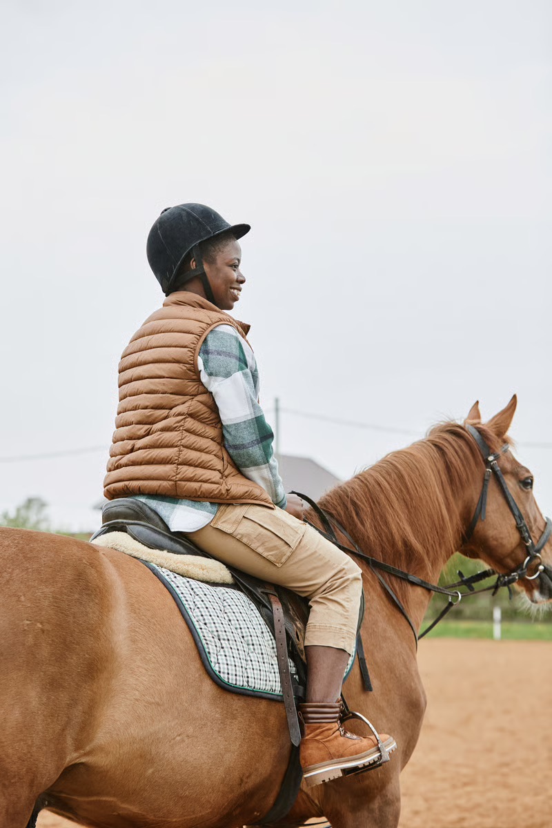 Black woman learning horseback riding while smiling on a horse during an outdoor riding lesson, celebrating Black equestrian culture and adult horsemanship.