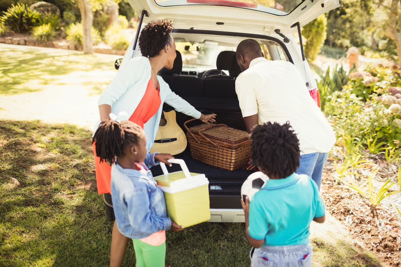 Black family packing a car for a summer road trip with a cooler and picnic basket before an outdoor adventure